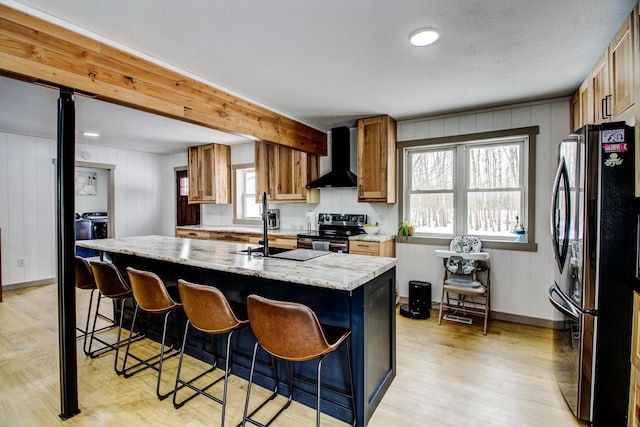 kitchen with an island with sink, stainless steel appliances, light stone countertops, light hardwood / wood-style floors, and wall chimney range hood