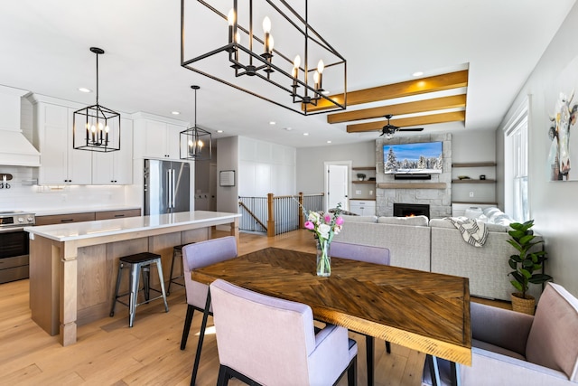 dining space featuring ceiling fan with notable chandelier, light hardwood / wood-style flooring, beam ceiling, and a stone fireplace