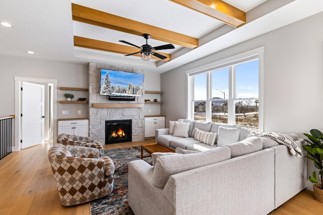 living room featuring a stone fireplace, beamed ceiling, a raised ceiling, ceiling fan, and light hardwood / wood-style flooring