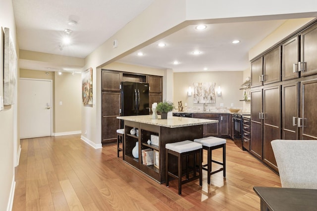 kitchen featuring a kitchen island, black appliances, a breakfast bar area, dark brown cabinetry, and light stone counters