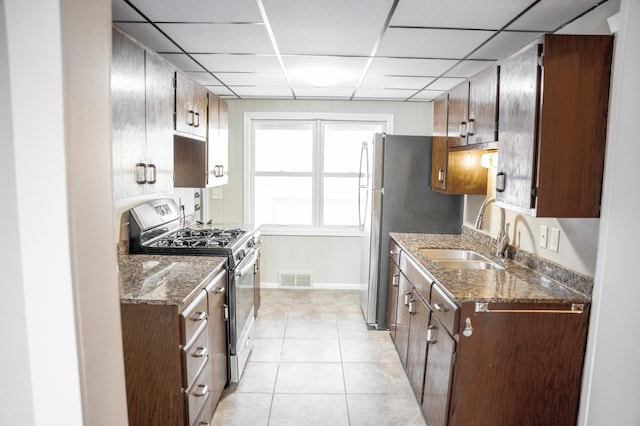 kitchen featuring light tile patterned floors, a drop ceiling, sink, stainless steel range with gas stovetop, and dark stone counters