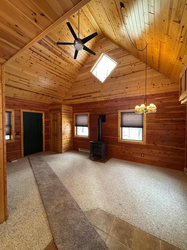 unfurnished living room featuring wood walls, wooden ceiling, carpet, a wood stove, and ceiling fan with notable chandelier