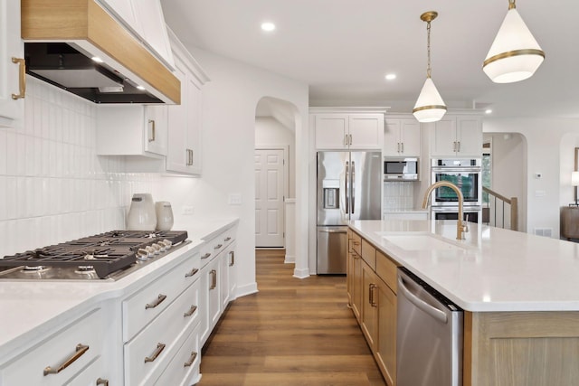 kitchen featuring custom exhaust hood, appliances with stainless steel finishes, hanging light fixtures, an island with sink, and white cabinetry