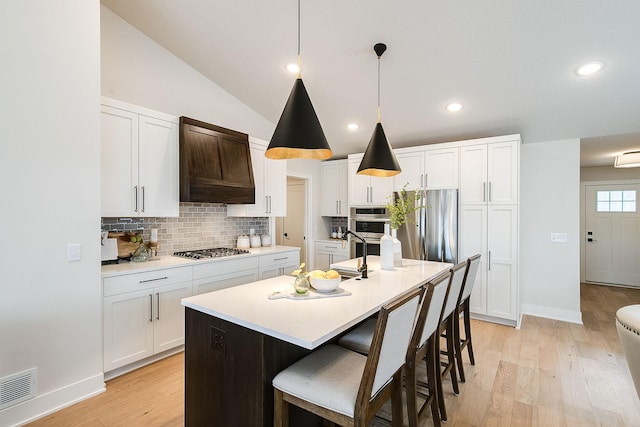 kitchen with pendant lighting, vaulted ceiling, an island with sink, appliances with stainless steel finishes, and white cabinetry