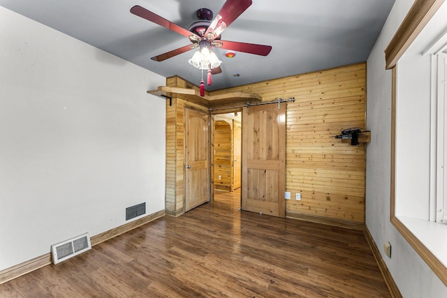 unfurnished bedroom with ceiling fan, a barn door, wooden walls, and dark hardwood / wood-style floors