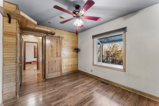 unfurnished bedroom featuring ceiling fan, a barn door, wooden walls, and dark hardwood / wood-style floors