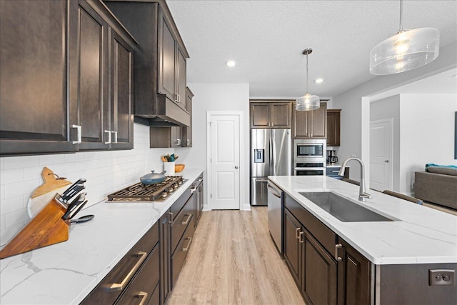 kitchen with an island with sink, a sink, dark brown cabinetry, light wood-style floors, and appliances with stainless steel finishes