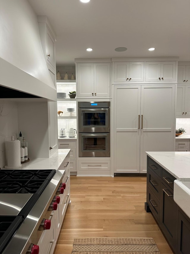 kitchen with white cabinetry, double oven, custom exhaust hood, and light hardwood / wood-style flooring