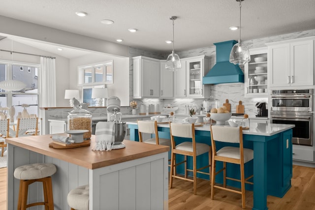 kitchen with double oven, premium range hood, white cabinets, backsplash, and a kitchen island