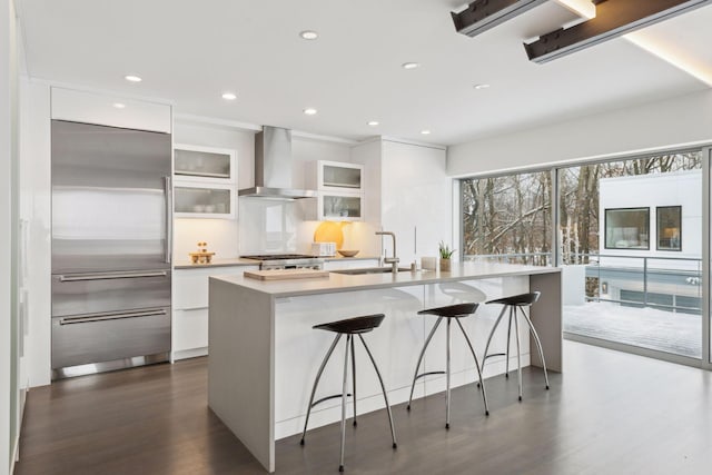 kitchen with a kitchen island with sink, stainless steel built in fridge, a kitchen bar, wall chimney exhaust hood, and white cabinetry