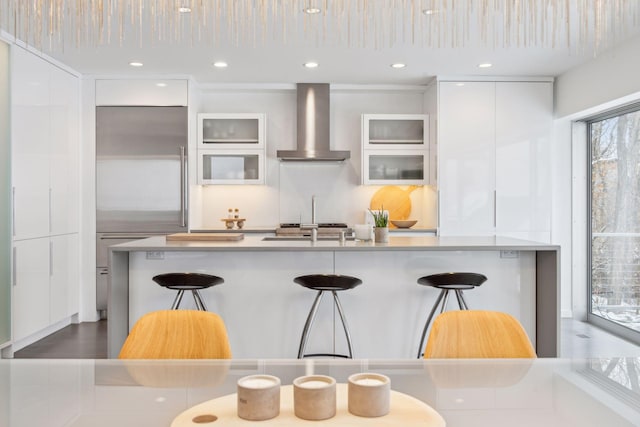 kitchen with a kitchen bar, stainless steel built in refrigerator, wall chimney range hood, and white cabinetry