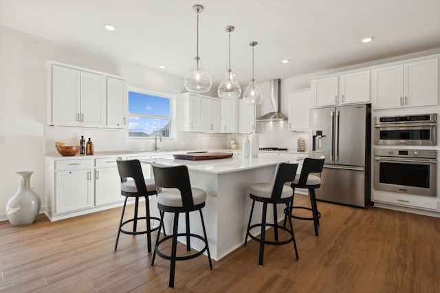 kitchen featuring appliances with stainless steel finishes, wall chimney range hood, decorative light fixtures, light hardwood / wood-style flooring, and white cabinets