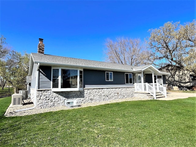 back of house with a shingled roof, a yard, stone siding, and a chimney