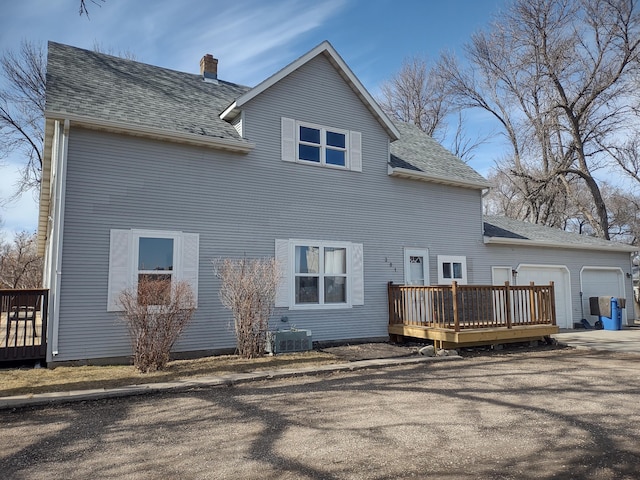 back of house with a wooden deck, an attached garage, a chimney, driveway, and a shingled roof