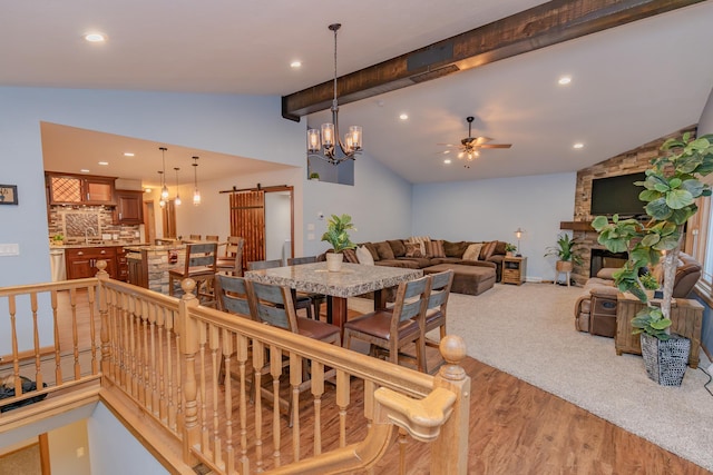 dining space featuring ceiling fan with notable chandelier, hardwood / wood-style floors, lofted ceiling with beams, a stone fireplace, and a barn door