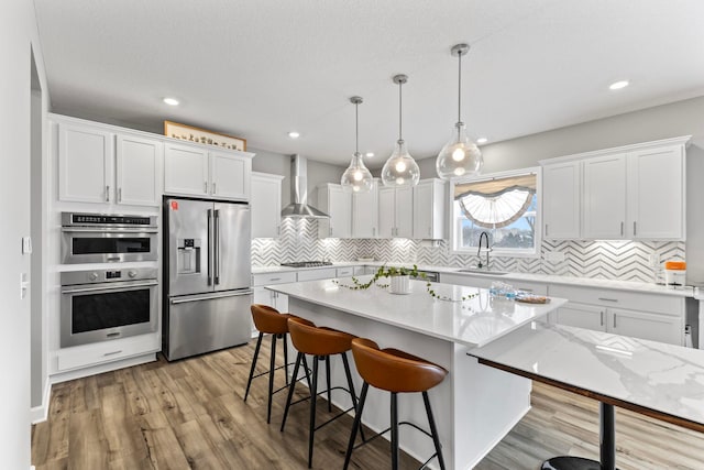 kitchen featuring a center island, decorative light fixtures, wall chimney range hood, white cabinetry, and appliances with stainless steel finishes