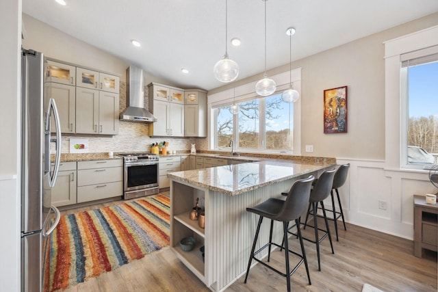 kitchen featuring appliances with stainless steel finishes, decorative light fixtures, light stone counters, kitchen peninsula, and wall chimney exhaust hood