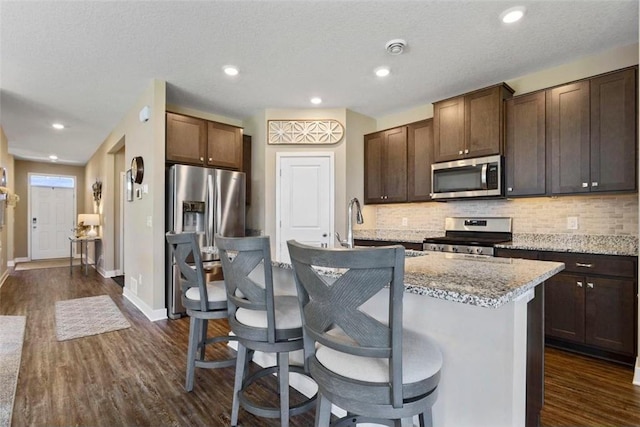 kitchen featuring appliances with stainless steel finishes, light stone countertops, dark hardwood / wood-style floors, a center island with sink, and dark brown cabinetry