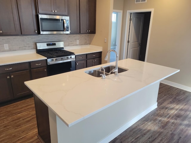 kitchen with stainless steel appliances, decorative backsplash, sink, light stone counters, and a center island with sink