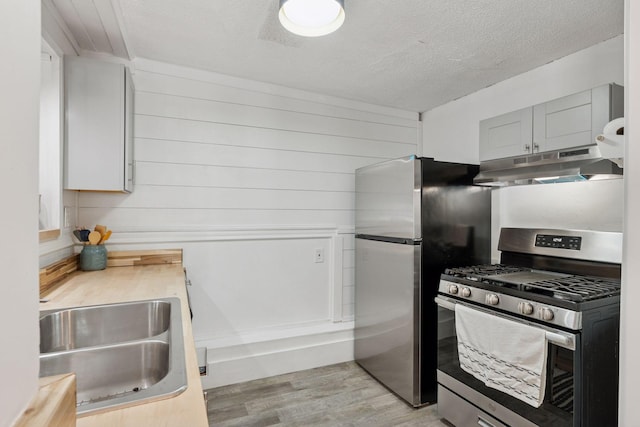 kitchen with a sink, stainless steel appliances, under cabinet range hood, a textured ceiling, and light wood-type flooring