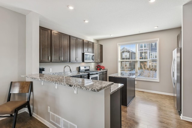 kitchen featuring light stone countertops, dark brown cabinetry, appliances with stainless steel finishes, kitchen peninsula, and a breakfast bar