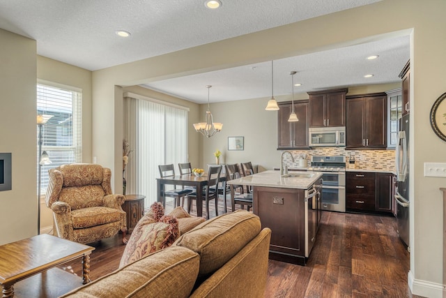 kitchen with light stone counters, decorative light fixtures, dark brown cabinets, stainless steel appliances, and a kitchen island with sink