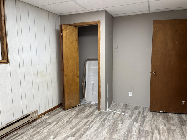 unfurnished bedroom with a paneled ceiling, wood walls, a baseboard radiator, and light hardwood / wood-style floors