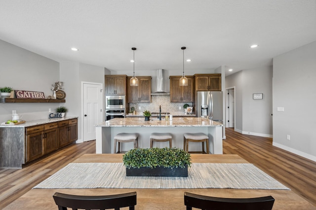 kitchen featuring appliances with stainless steel finishes, a breakfast bar, decorative light fixtures, a kitchen island with sink, and wall chimney exhaust hood