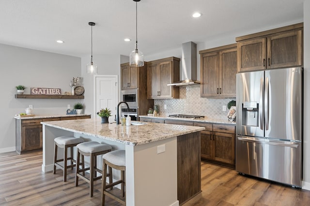 kitchen featuring sink, appliances with stainless steel finishes, light stone countertops, a center island with sink, and wall chimney exhaust hood