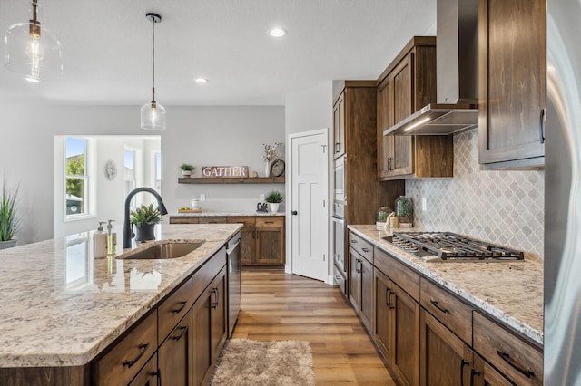 kitchen featuring an island with sink, sink, light stone counters, stainless steel appliances, and wall chimney exhaust hood