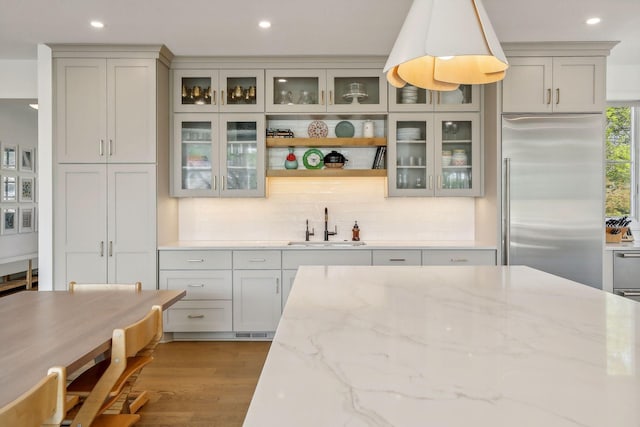 kitchen with light stone countertops, pendant lighting, wood-type flooring, stainless steel built in fridge, and sink