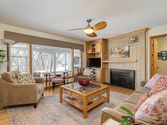 living area featuring light wood-style floors, a brick fireplace, ceiling fan, and a textured ceiling