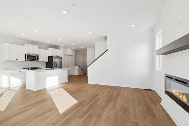 kitchen with white cabinetry, stainless steel appliances, a center island with sink, decorative backsplash, and light wood-type flooring