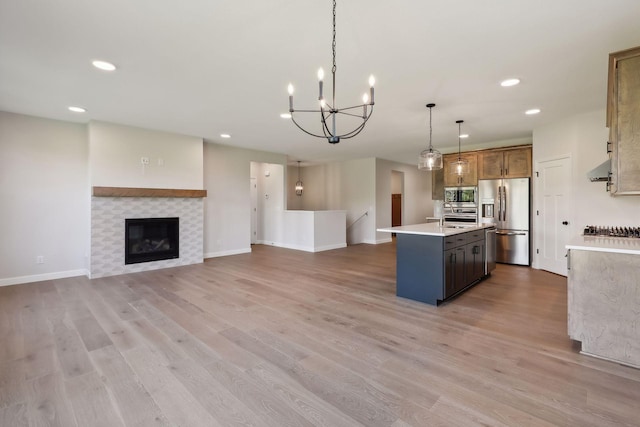 kitchen with light hardwood / wood-style flooring, stainless steel appliances, a fireplace, hanging light fixtures, and a center island with sink