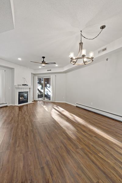 unfurnished living room featuring wood-type flooring, a textured ceiling, a baseboard radiator, a tile fireplace, and ceiling fan with notable chandelier