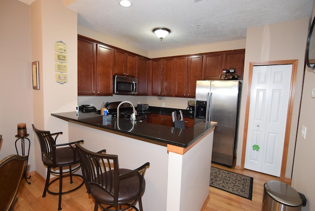 kitchen with kitchen peninsula, a textured ceiling, stainless steel appliances, light hardwood / wood-style floors, and a breakfast bar