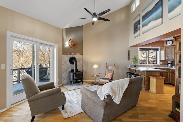 living room featuring ceiling fan, baseboards, a wood stove, light wood-style floors, and a towering ceiling