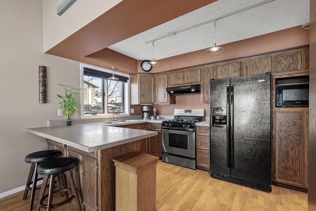 kitchen featuring under cabinet range hood, a peninsula, light wood-style floors, black appliances, and a sink