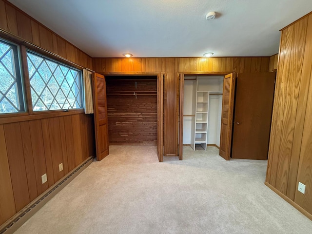 unfurnished bedroom with a baseboard radiator, two closets, light colored carpet, and wood walls