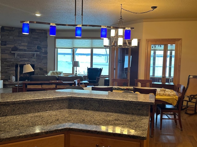 kitchen with a textured ceiling, wood-type flooring, a stone fireplace, hanging light fixtures, and crown molding