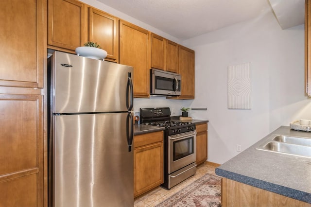 kitchen with light tile patterned floors, a sink, appliances with stainless steel finishes, brown cabinets, and dark countertops