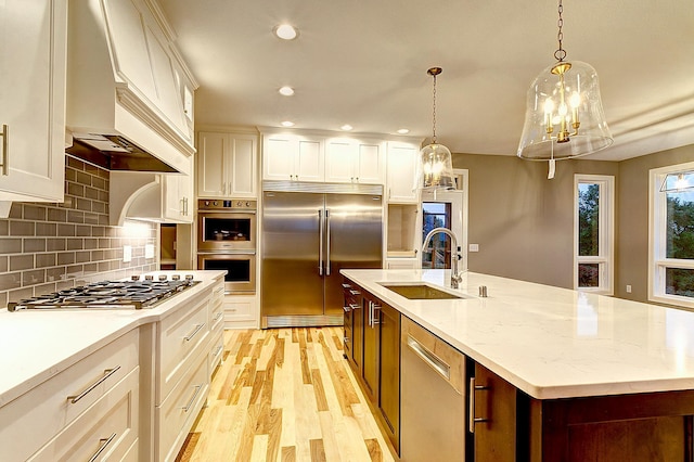 kitchen with custom exhaust hood, stainless steel appliances, an island with sink, sink, and hanging light fixtures