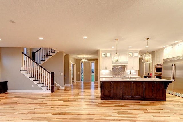 kitchen featuring appliances with stainless steel finishes, hanging light fixtures, a kitchen island with sink, decorative backsplash, and light hardwood / wood-style flooring