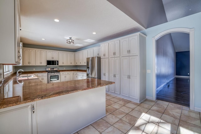 kitchen featuring arched walkways, stainless steel appliances, a peninsula, a sink, and dark stone countertops