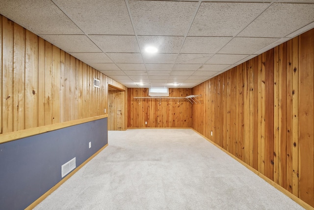 basement featuring carpet flooring, a wall mounted AC, a drop ceiling, and wood walls