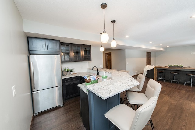 kitchen with a breakfast bar area, dark wood-type flooring, hanging light fixtures, light stone countertops, and stainless steel refrigerator