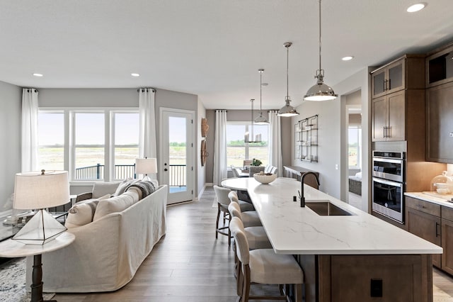 kitchen with an island with sink, stainless steel double oven, pendant lighting, light stone counters, and sink