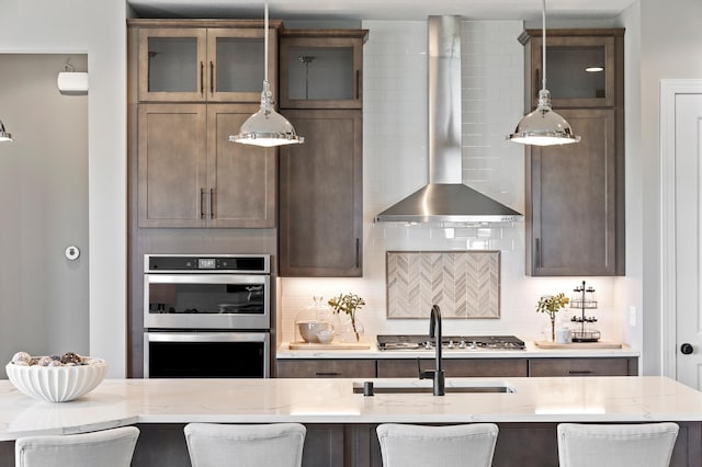 kitchen with light stone countertops, wall chimney exhaust hood, stainless steel appliances, decorative backsplash, and a kitchen breakfast bar