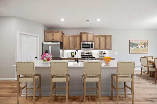 kitchen with light wood-type flooring, brown cabinets, a kitchen island with sink, a sink, and appliances with stainless steel finishes