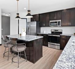 kitchen with decorative light fixtures, stainless steel appliances, light stone counters, dark brown cabinets, and a breakfast bar area